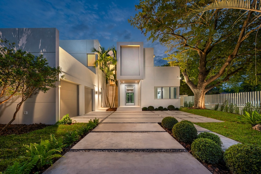 <span class="uk-h4">Two-story luxury home at dusk with warm exterior lighting, a concrete paver driveway, and palm trees.</span>