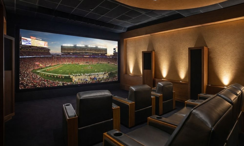<span class="uk-h4">Home theater with leather reclining seats and in-wall speakers, large screen showing a live football broadcast.</span>