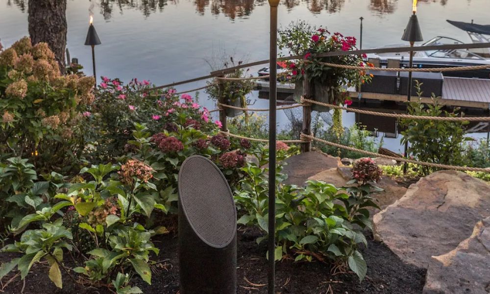 <span class="uk-h4">Coastal Source outdoor speaker installed in a garden area with a boat dock in the background.</span>