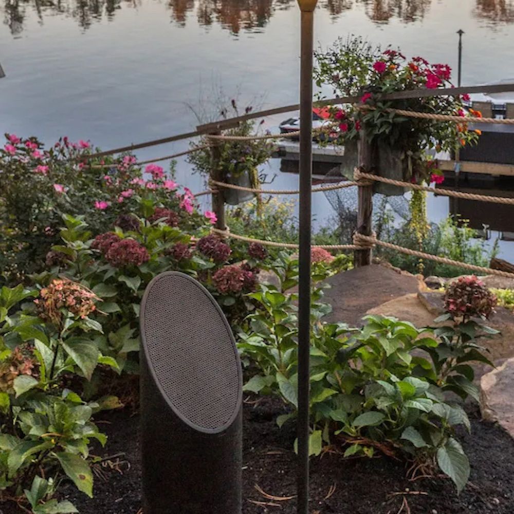 <span class="uk-h4">Coastal Source outdoor speaker installed in a garden area with a boat dock in the background.</span>