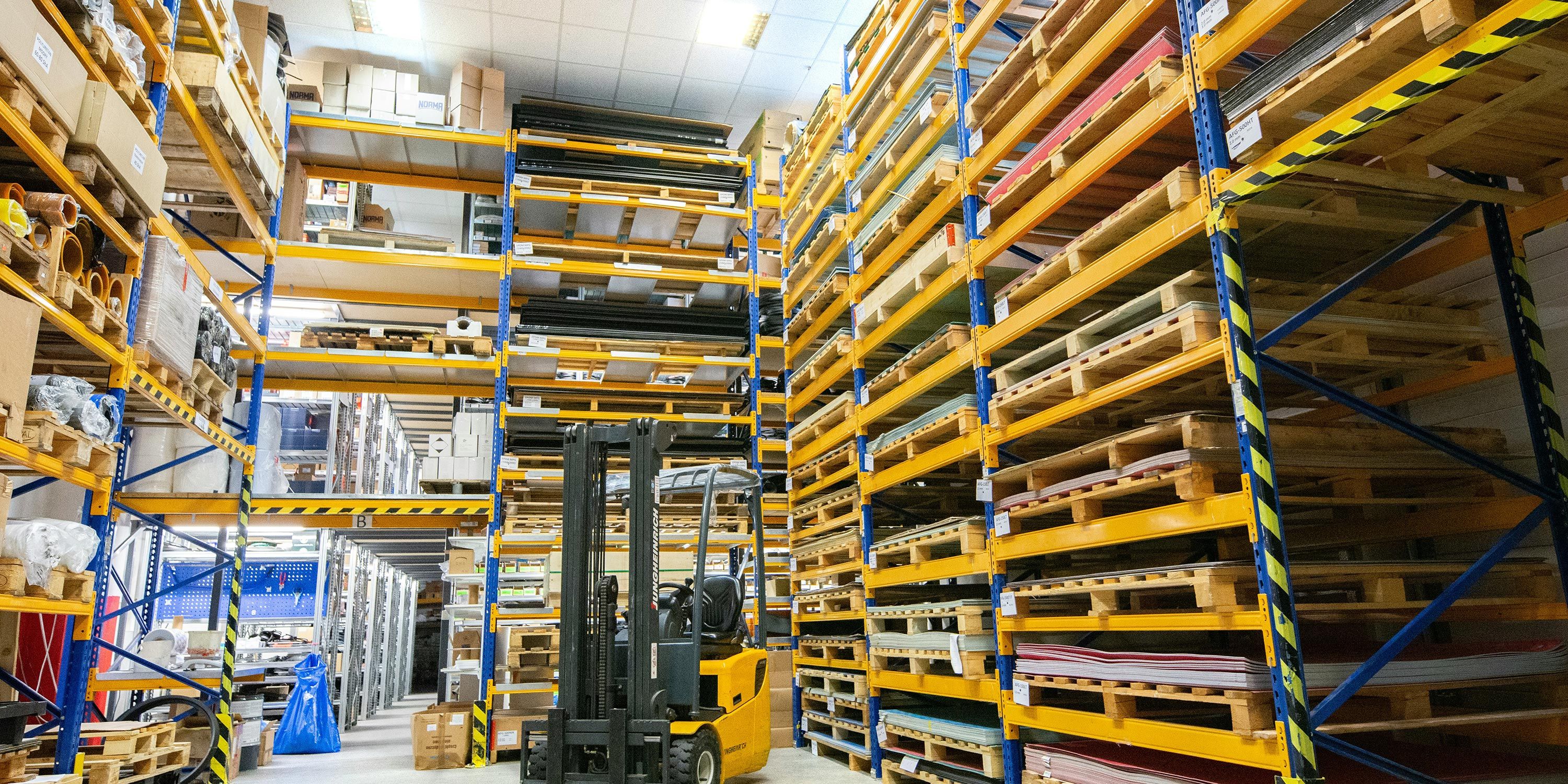 A well-organized industrial warehouse with tall yellow shelving, stocked pallets, and a forklift in the aisle.