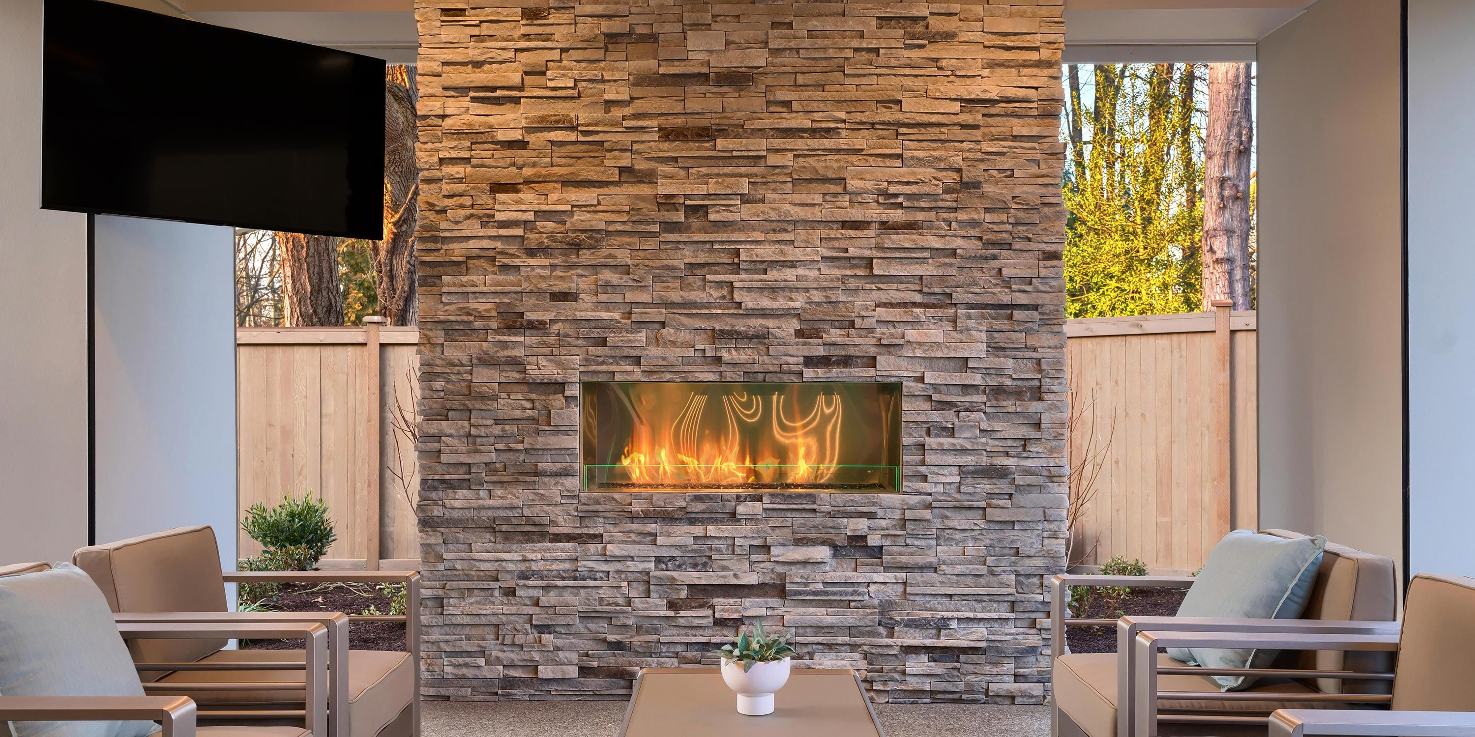 A cozy outdoor space with a modern stone fireplace, cushioned seating, and a wooden privacy fence.