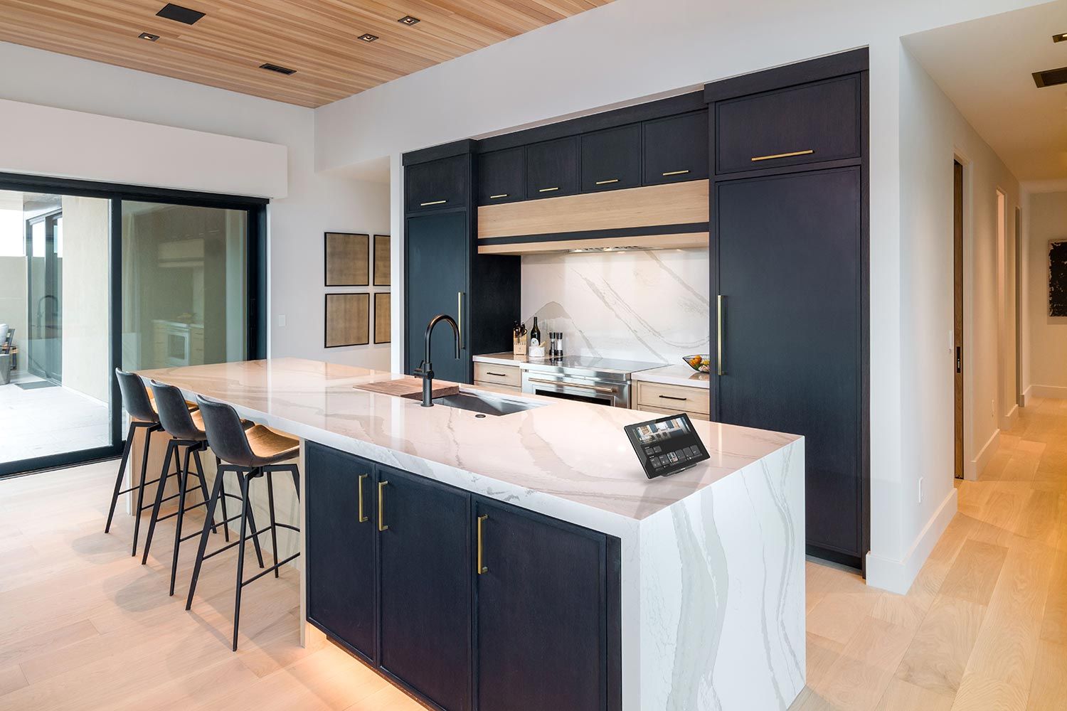 A modern kitchen with a large marble island, black cabinets, and a tablet displaying automation controls.