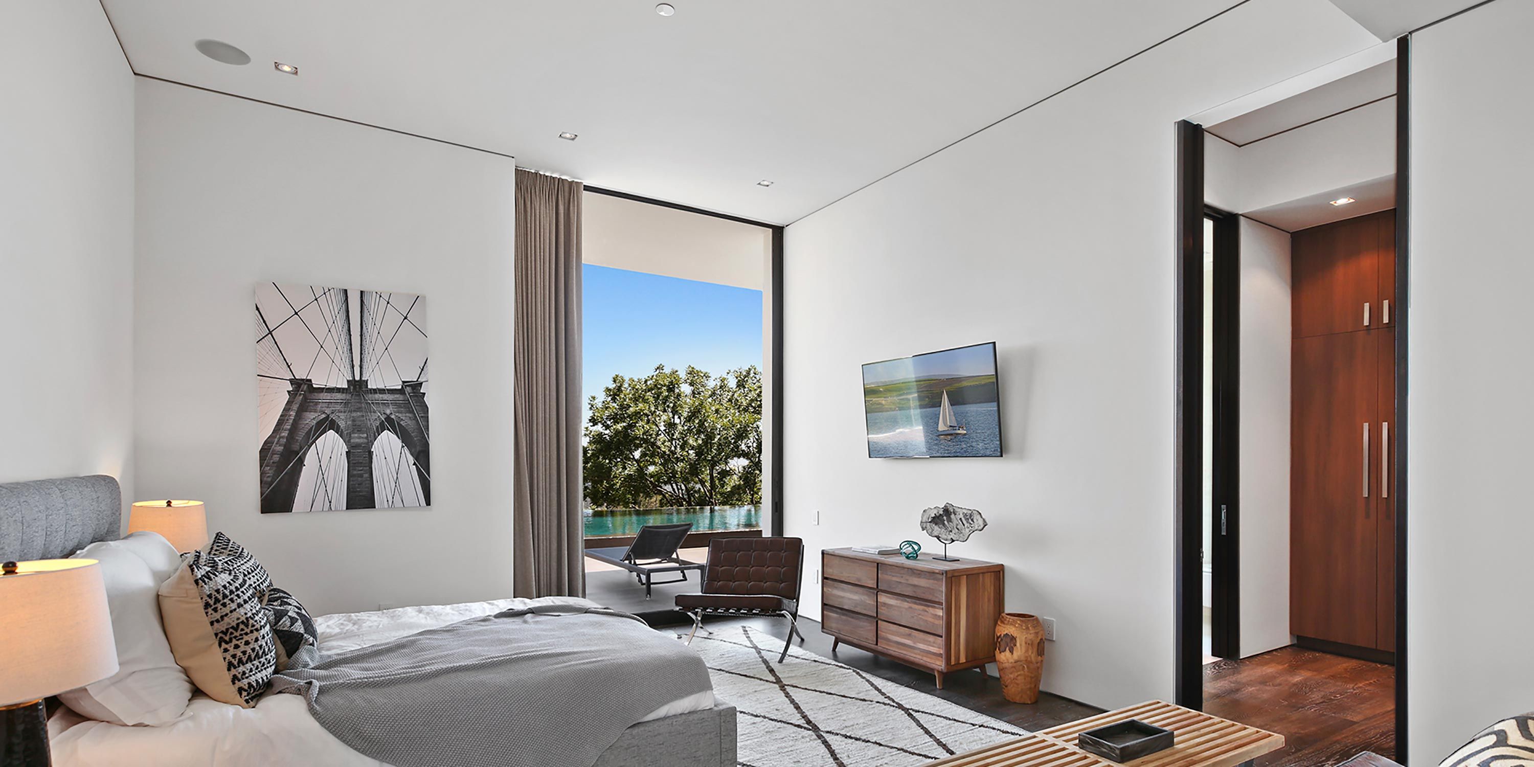 Modern bedroom with large windows, minimal decor, and a serene view of a pool and greenery.