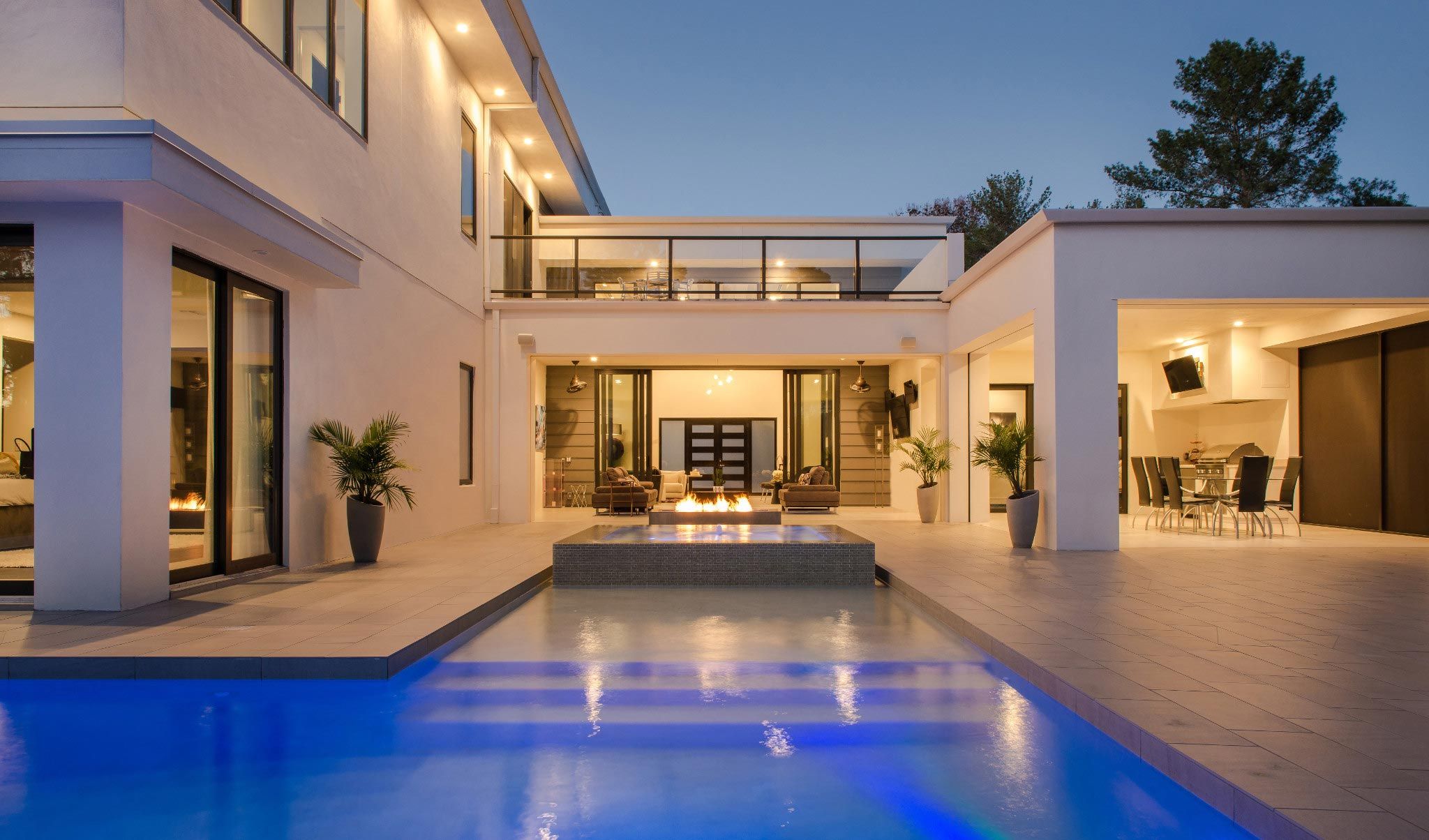 Modern luxury home exterior at dusk with a glowing swimming pool, fire feature, and spacious patio.