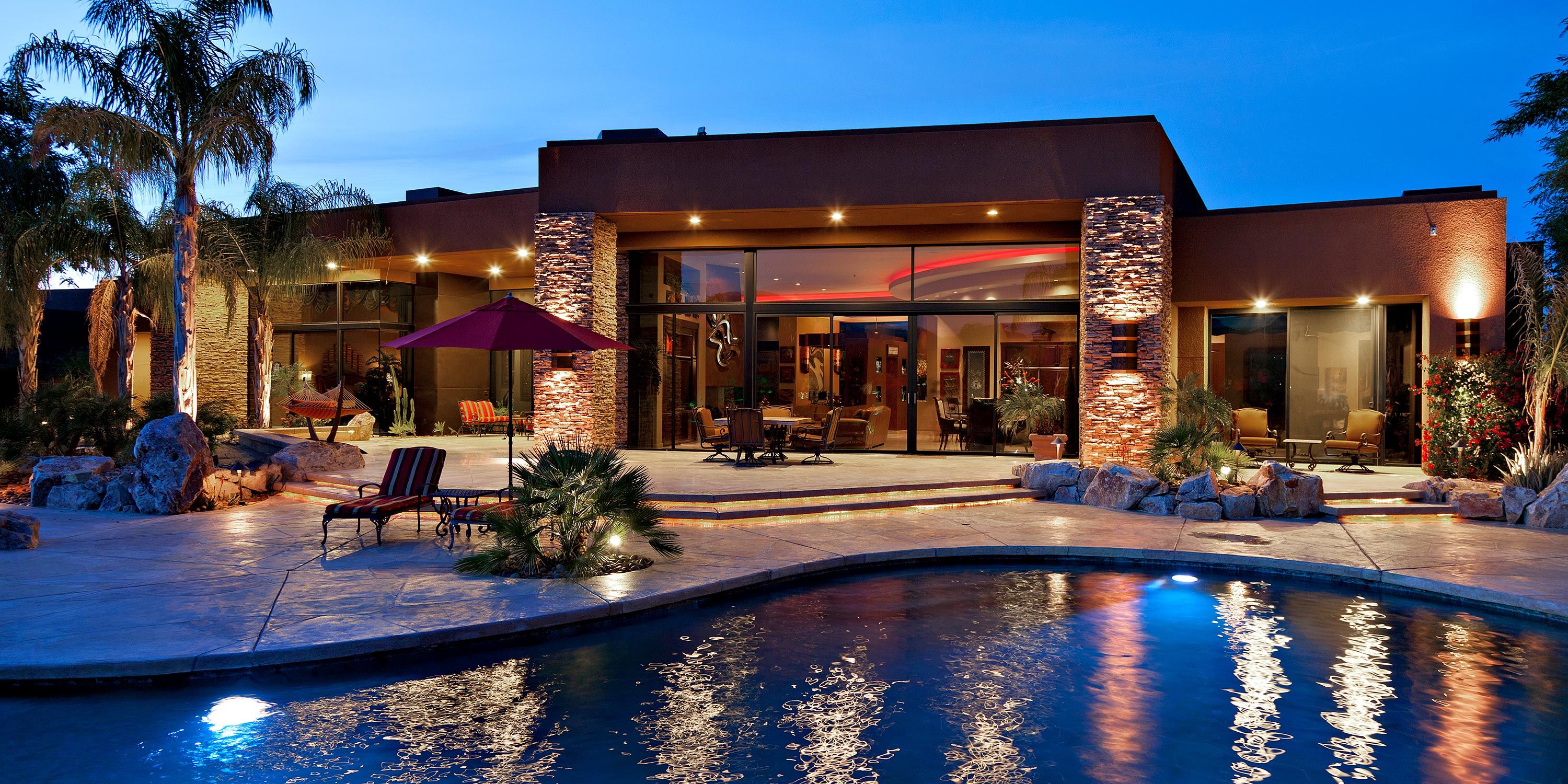 A luxurious backyard with a swimming pool, spa, and illuminated patio at twilight.