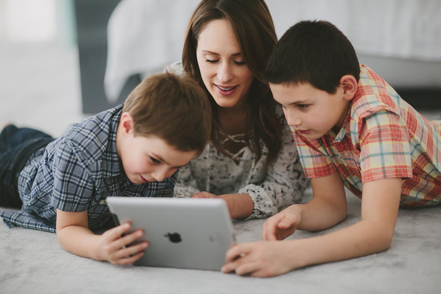 Mother and two kids laying on the floor, enjoying a tablet together in a cozy setting.